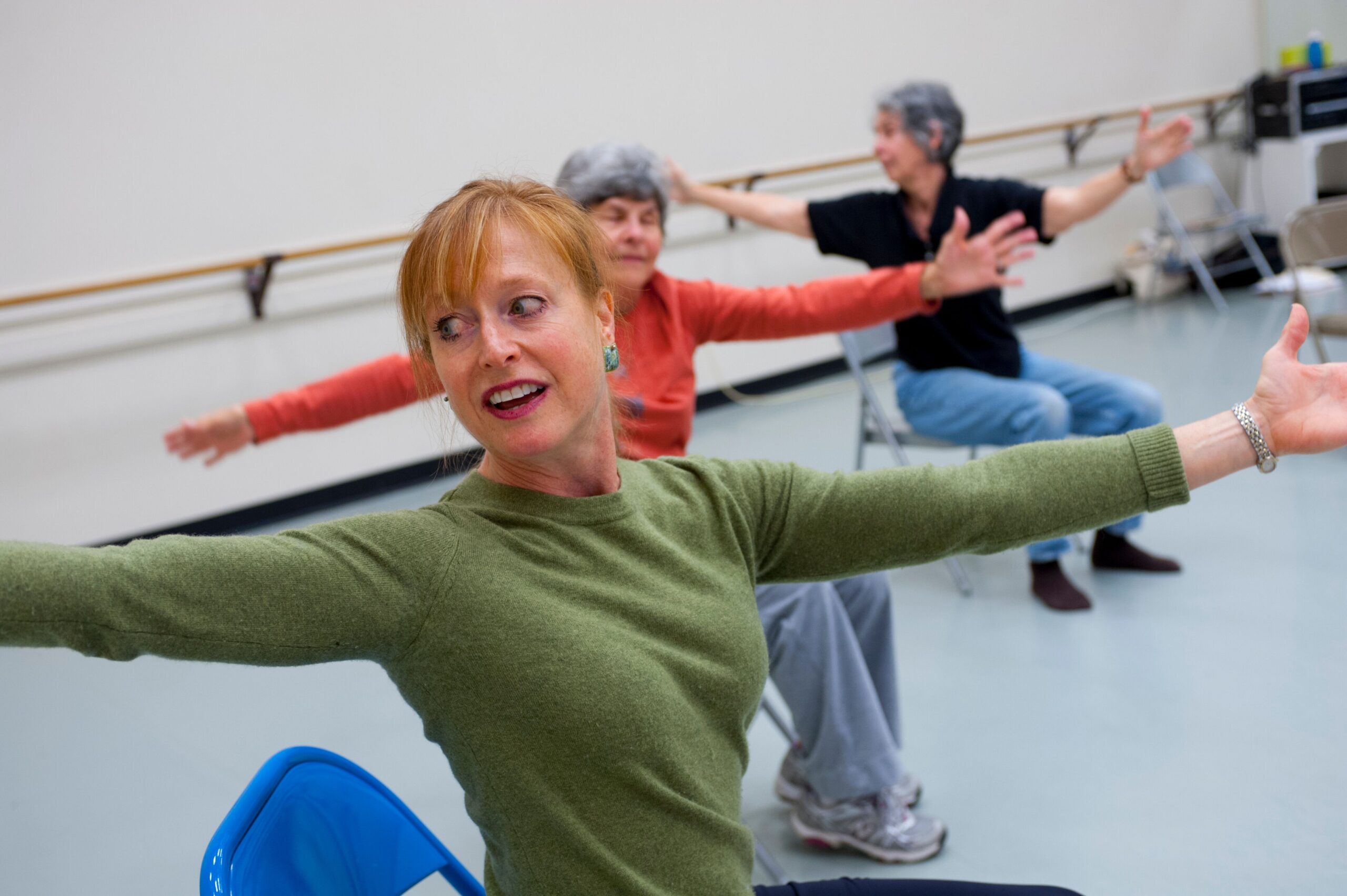 Dance teacher in green sweater leads a seated dance activity.